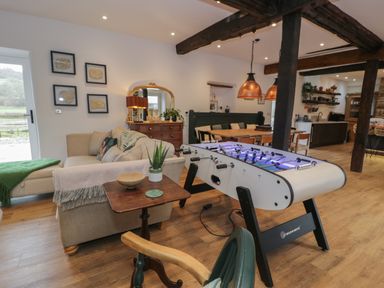 A living area with a foosball table, sofa, wooden side table, dining table with bench, and kitchen shelves at Plas Glan Yr Afon in Llangernyw