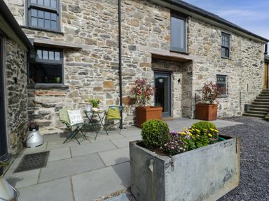 An outdoor stone patio with metal chairs and table and large planters in front of a stone building at Plas Glan Yr Afon in Llangernyw