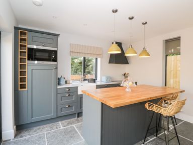 A kitchen with grey cabinets wooden island with wicker stools and three hanging yellow lights at Little Lugg in Cross Keys near Hereford