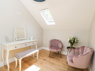 A room with a dressing table with mirror and bench and two pink upholstered chairs and a small round table with flowers at Little Lugg Cross Keys near Hereford