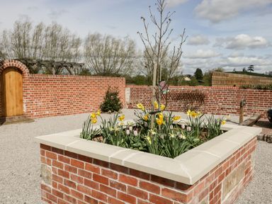 A garden area with a square raised brick flower bed containing daffodils and a small tree and a wooden door set in a brick wall at Little Lugg in Cross Keys near Hereford