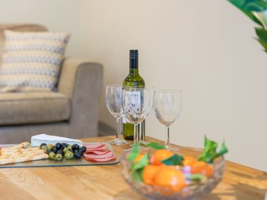 A table with wine glasses a bottle of wine a bowl of fruit and a platter of cheese olives and sliced meat in a living room with a couch and cushion at Golden Sands in Carbis Bay