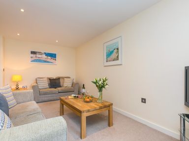 A living room with two sofas a wooden coffee table with flowers and snacks and framed pictures on the walls at Golden Sands in Carbis Bay