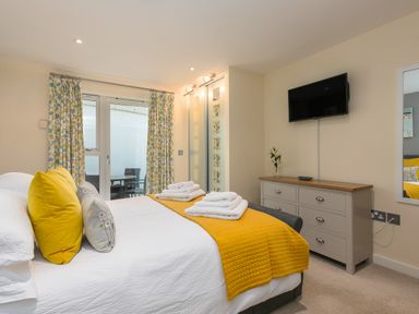A bedroom with a bed with yellow and gray pillows a yellow blanket and folded towels a dresser with a vase and a wall mounted TV at Golden Sands in Carbis Bay