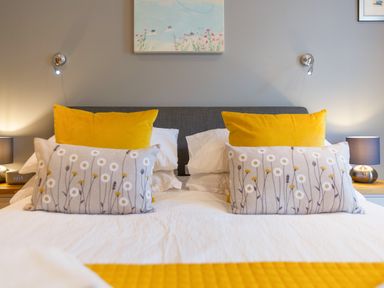 A bed with yellow and floral pillows bedside tables with lamps and a wall painting above the headboard at Golden Sands in Carbis Bay