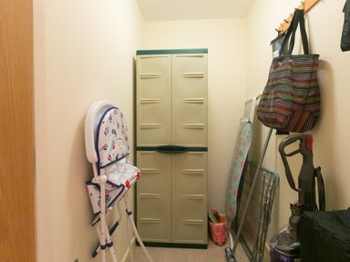 A storage room with a closed cabinet a folded high chair an ironing board cleaning tools and bags at Golden Sands in Carbis Bay