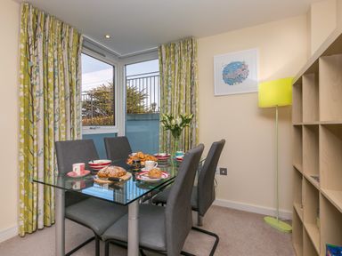 A dining room with a glass table set for breakfast with pastries and red and white bowls and cups and grey chairs with floral curtains and a floor lamp at Golden Sands in Carbis Bay