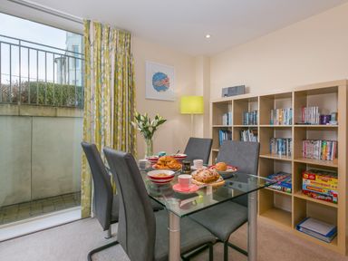 A dining room with a glass table and four chairs next to a bookshelf with books and games at Golden Sands in Carbis Bay