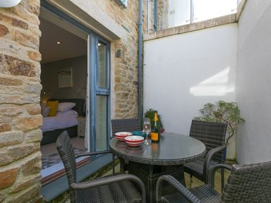 An outdoor patio area with a round glass table and four wicker chairs next to a stone wall and an open door leading to a bedroom at Golden Sands in Carbis Bay