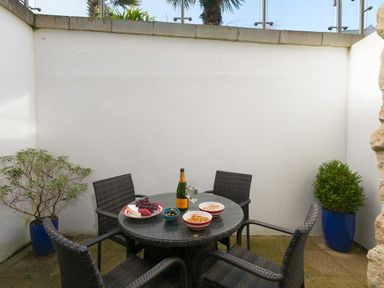 An outdoor patio with a round table and four wicker chairs a bottle of champagne two glasses and snacks on the table with potted plants in blue containers at Golden Sands in Carbis Bay