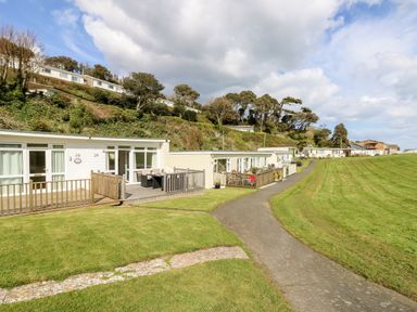A row of single-story holiday homes with wooden decks along a paved pathway and a grassy hill with trees in the background at Seas The Day in Ventnor