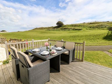 An outdoor dining area with a wicker table and chairs set on a wooden deck overlooking a grassy hill at Seas The Day in Ventnor