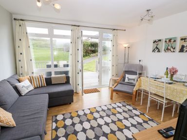 A living room with a gray sofa, an armchair, a dining table with chairs, a patterned rug, and large windows with curtains at Seas The Day in Ventnor