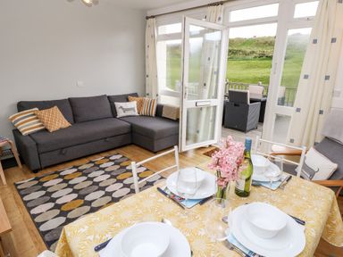 A living room with a gray sofa cushions on a geometric rug and a dining table set with white dishes flowers and wine at Seas The Day in Ventnor