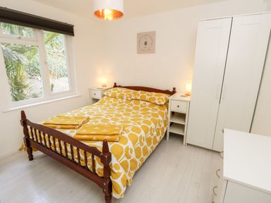 A bedroom with a wooden bed with yellow patterned bedding two white nightstands with lamps a white wardrobe and a window showing green plants at Seas The Day in Ventnor