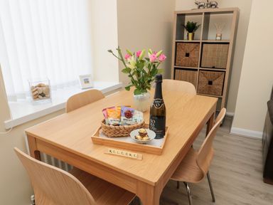 A dining room with a wooden table and chairs a vase with flowers a basket of snacks a bottle of wine and a storage unit with baskets at Stones Throw in Amble