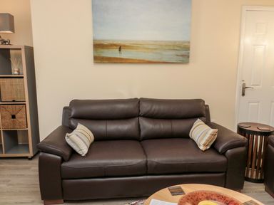 A brown leather sofa with two striped cushions a wooden side table a shelving unit and a painting on the wall at Stone's Throw in Amble