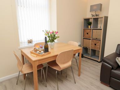 A dining area with a wooden table and four chairs a vase with flowers a bottle and snacks on the table a shelving unit with baskets and a lamp at Stones Throw in Amble