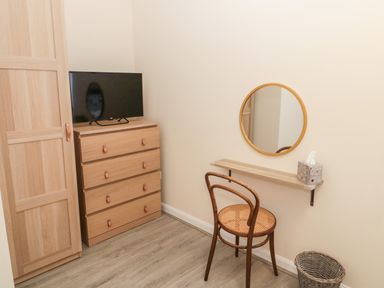 A corner of a room with a wooden wardrobe and chest of drawers with a TV on top a round wall mirror above a small shelf with a chair and a wicker basket on the floor at Stone's Throw in Amble
