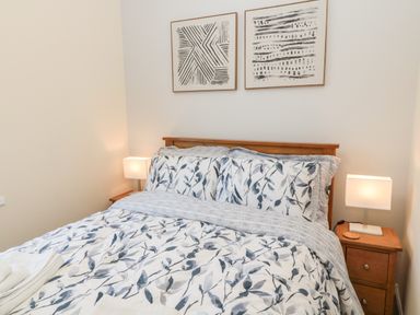 A bedroom with a bed with leaf patterned bedding two wooden bedside tables with lamps and abstract wall art at Stone's Throw in Amble