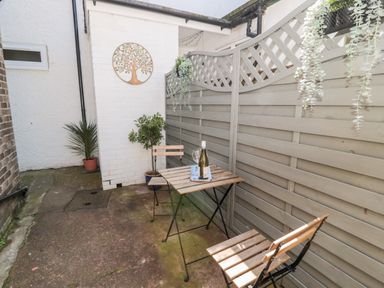 An outdoor patio with a small wooden table and two chairs a bottle of white wine and glass on the table potted plants and hanging greenery on a fence at Stone's Throw in Amble