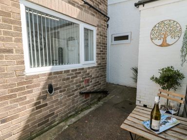 An outdoor narrow patio area with a small wooden table holding a bottle of wine and two wine glasses and a metal tree wall decoration at Stone's Throw in Amble