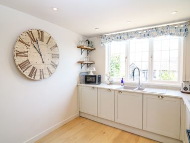 A kitchen with a large wall clock wooden shelves and a window with patterned curtains at Hillside in Crafthole