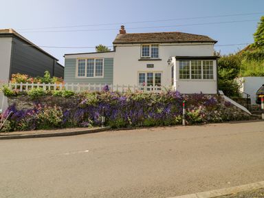 A white cottage with blue siding and a flower garden in front along a road at Hillside in Crafthole