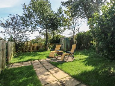 A garden with two chairs and a fire pit on a paved area surrounded by grass and trees at Hillside in Crafthole