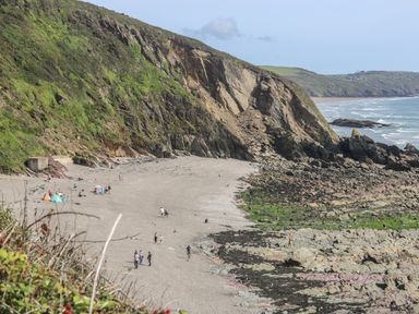 A sandy beach with people walking and sitting near cliffs and rocky shoreline at Hillside in Crafthole