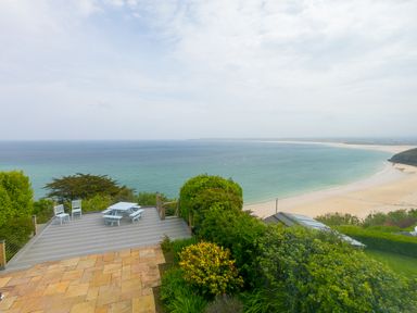 A terrace with white chairs and a table overlooking a curved sandy beach and ocean at Moonbeams in Carbis Bay