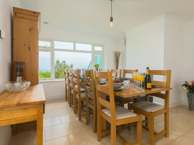 A dining room with a wooden table set for a meal wooden chairs a sideboard and a large window at Moonbeams in Carbis Bay