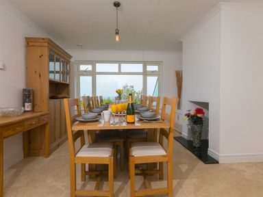 A dining room with a wooden table set for eight near large windows at Moonbeams in Carbis Bay