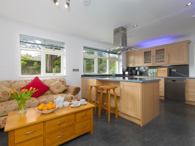 A kitchen with wooden cabinets an island with stools a sofa and a wooden coffee table with fruit and tea set at Moonbeams in Carbis Bay