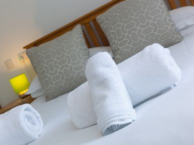 A bed with white rolled towels and patterned cushions with a wooden headboard and a bedside table with a green lamp at Moonbeams in Carbis Bay