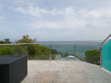 A balcony with stone tile flooring and glass railing overlooking the sea with outdoor furniture below at Moonbeams in Carbis Bay