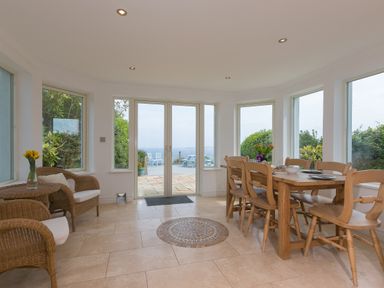 A dining area with a wooden table and chairs a small table with wicker chairs and flowers outside doors leading to a patio at Moonbeams in Carbis Bay