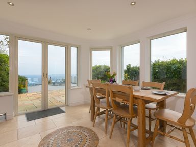 A dining area with a wooden table and six chairs near large windows and glass doors overlooking an outdoor patio and garden at Moonbeams in Carbis Bay