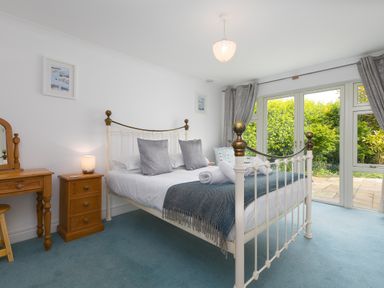 A bedroom with a white metal bed frame gray pillows and blue blanket next to wooden bedside table and dressing table with window curtains at Moonbeams in Carbis Bay