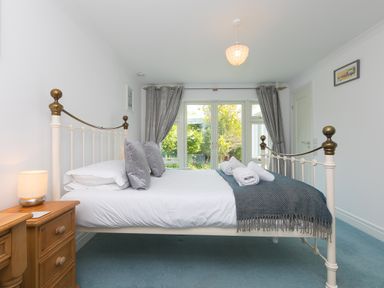 A bedroom with a metal bed frame white bedding pillows and rolled towels on a blue throw next to wooden bedside tables with a lamp and garden view through glass doors at Moonbeams in Carbis Bay