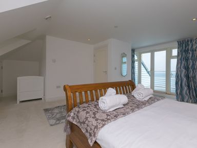 A bedroom with a wooden bed with rolled towels on it a small white chest a floor rug and large windows with curtains at Moonbeams in Carbis Bay