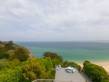 A deck with white outdoor furniture overlooking the ocean and a sandy beach surrounded by trees at Moonbeams in Carbis Bay