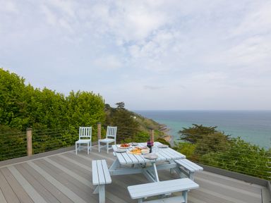 An outdoor deck with a picnic table set for food and two chairs overlooking the sea and trees at Moonbeams in Carbis Bay