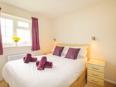 A bedroom with a double bed purple pillows and towels bedside drawers and a window with purple curtains at West Lulworth Apartment in West Lulworth