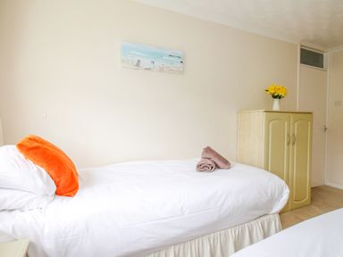 A bedroom with a single bed with orange pillow and towels on it a cabinet with flowers on top and a door at West Lulworth Apartment in West Lulworth