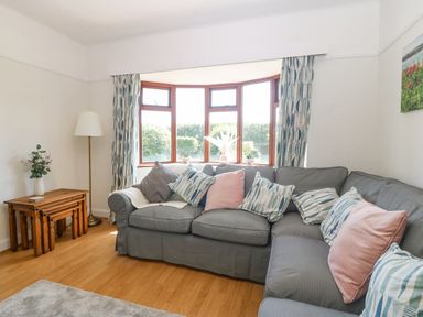 A living room with a gray sectional sofa and patterned cushions near a bay window with blue patterned curtains and nested wooden tables at Awelfryn in Elim near Llanddeusant