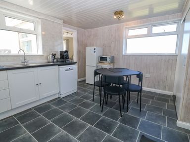 A kitchen with white cabinets a dishwasher a refrigerator a microwave on a small table a black dining table with four chairs and two large windows at Awelfryn in Elim near Llanddeusant