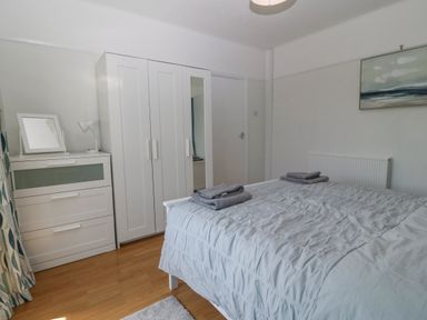 A bedroom with a bed covered with a grey comforter and towels a white wardrobe and chest of drawers with a lamp and mirror a painting on the wall and patterned curtains at Awelfryn in Elim near Llanddeusant