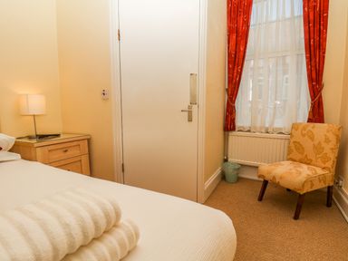 A bedroom with a bed with folded towels a wooden nightstand with a lamp an upholstered chair next to a window with red curtains at Lonsdale Villa Scarborough