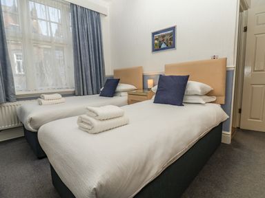 A bedroom with two single beds with white bedding blue pillows folded towels nightstand with lamp and window with blue curtains at Lonsdale Villa in Scarborough